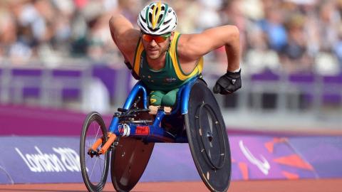 Wheelchair racer Kurt Fearnley is captured racing at the London 2012 Paralympics. A packed stadium is blurred behind him.