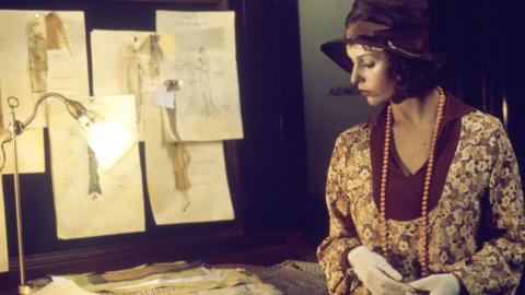 Film still of Helen Morse as Caddie looking at drawings of women's dress designs in a studio.
