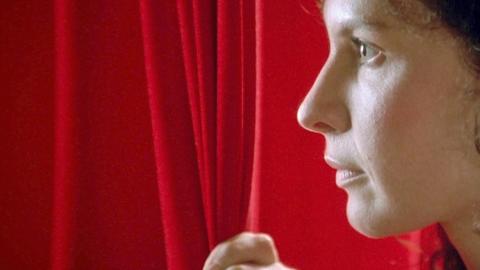 Close up of Tara Morice in profile as her character Fran in the film Strictly Ballroom. Fran his pulling aside a red curtain and looking past it, facing left of camera.