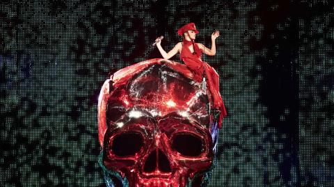 Kylie Minogue, dressed entirely in red, is sitting on top of a giant red shiny skull prop at one of her live shows. 