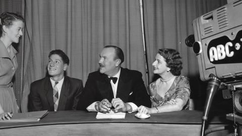 Four people are in a studio setting. Three of them sit behind a desk and the fourth stands. They are all looking at her. There is a film camera pointed at them that says ABC TV on the side.