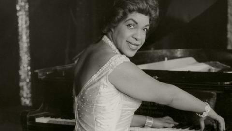 Winifred Atwell seated at the piano turns to smile at the camera