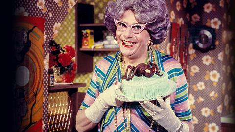 Dame Edna Everage is in a very colourful kitchen. She (Barry Humphries in drag) is holding an iced cake with a chocolate spider on top. She has wisteria-coloured hair and sparkling pink glasses.