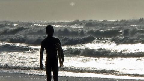 Silhouette of a man with his back to camera walking into the surf carrying a surfboard under one arm.