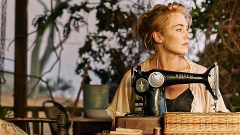 Kate Winslet in a scene from The Dressmaker. She is seated outside on a porch behind a table with a vintage singer sewing machine on top and looking sideways into the distance.