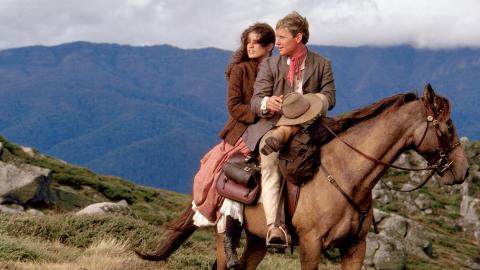 Sigrid Thornton and Tom Burlinson on a chestnut coloured horse with mountains in the background in a scene from 'The Man From Snowy River'.