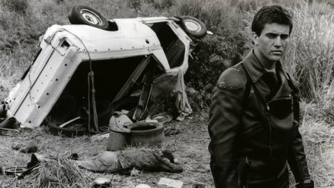 Mad Max dressed in his motorcycle cop leathers stands in front of an overturned car, looking dispassionately away