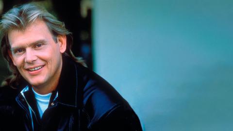 Singer John Farnham, circa late 1980s wearing a black jacket, pictured from the shoulders up. He has his head tilted slightly towards the camera and is smiling.