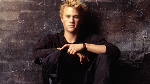 Heath Ledger is looking directly at the camera in a quietly confident pose. He is wearing black and up against a wall.