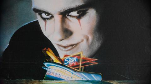 A close-up of a menacing looking man with dark eye make-up and an evil grin. His head looms over a smaller image of a neon-lit drive-in cinema and empty road.