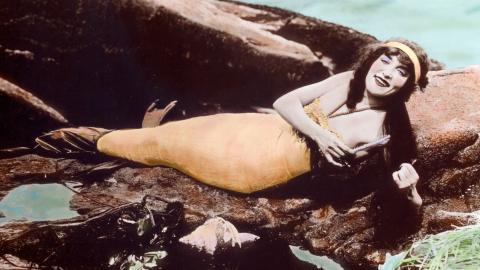 A hand-tinted photograph of Annette Kellerman wearing a mermaid costume and lying on rocks near the water.