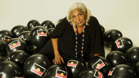 Trisha Morton-Thomas wearing a black dress and surrounded by black balloons with the Aboriginal flag on them