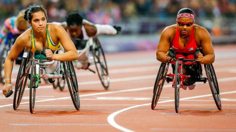 Women wheelchair athletes competing on an Olympic race track. There are two in the foreground who are side by side and another two can be seen further down the track.