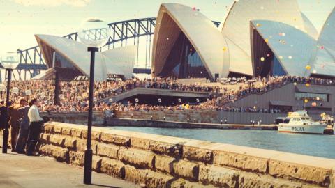 People line the boardwalk and steps of the Sydney Opera House for its opening. There are balloons in the air and the Sydney Harbour Bridge is in the background.