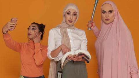 Three young women - two wearing the hijab - stand next to each other. One is taking a mobile phone selfie, another in corporate dress holds a law book, while the third wields a baseball bat.