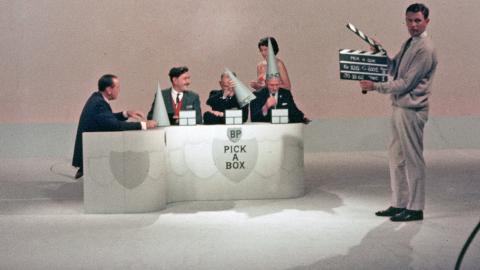 A group of people behind a desk in a TV studio with a man standing in front of them holding a clapper board. 