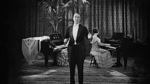 Jack O'Hagan singing. He is dressed in a suit. A woman accompanies him on the piano.