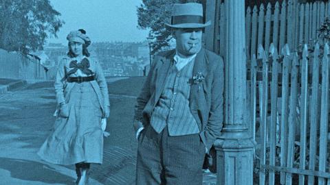 A scene from the film The Sentimental Block featuring Arthur Tauchert as 'The Bloke' leaning against a lamp post on a street corner Actress Lottie Lyell (Doreen) is walking up the street behind him.