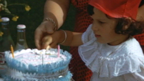 A little girl in a red party hat is looking at her birthday cake with candles on it and blue paper fringing around the outside of the cake.