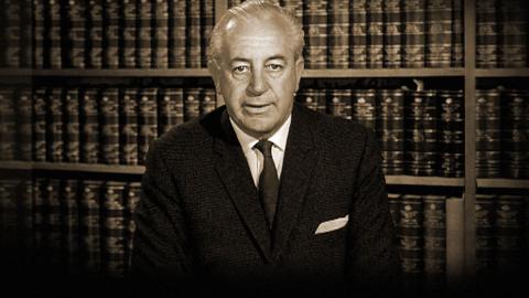 A sepia image of Harold Holt as federal treasurer, seated behind a desk and recording an address to the nation