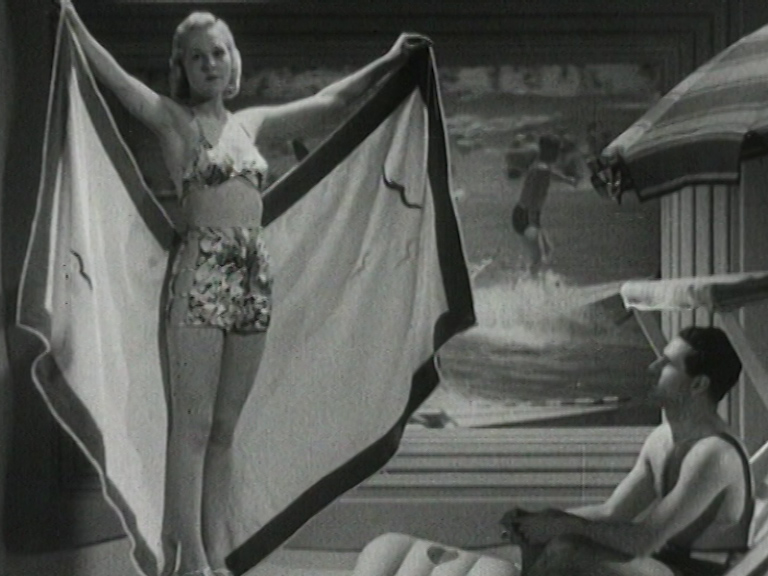 A model stands opening a towel to reveal her bikini to a man seated at the 'beach' during the fashion show